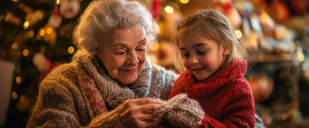 An elderly woman sitting in front of a christmas tree with her granddaughter looking at a gift and smiling.